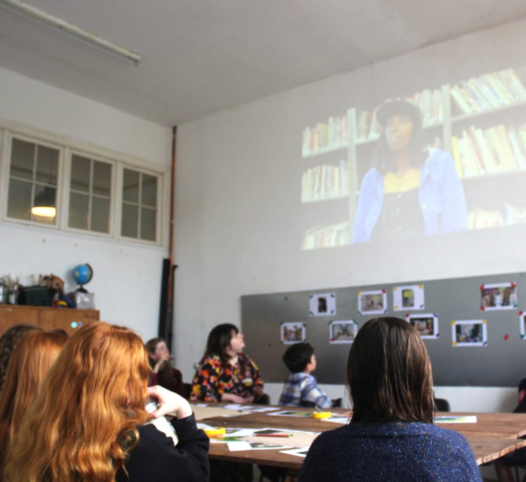 A group of young people sit watching a film projected on the wall.