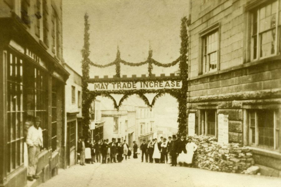 Archway erected in Helston to celebrate the start of construction of the railway to Helston, 22 March, 1882. The archway is in Church Street with the words "May Trade Increase" and "Progression". A group of people are standing underneath the archway and a man is standing in the doorway of Martyn's shop. The railway opened nearly 5 years later in May 1887