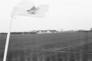 Black and white film photograph of an empty open grass football pitch, with white flag waving in the wind in the foreground and a single storey building in the background. 