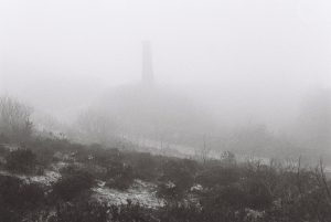 Black and white film photograph, showing the faint outline of a single industrial brick chimney in through the mist. 