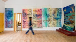 Blurred image of the artist walking past five large portrait format printed panels, (each bigger than a standard door), in bright shades with detail not discernible from this distance. 