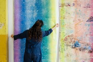 The artist positioning a large unstretched canvas, with vertical bands of colour fading into one another (purple, blue, turquoise, yellow). 