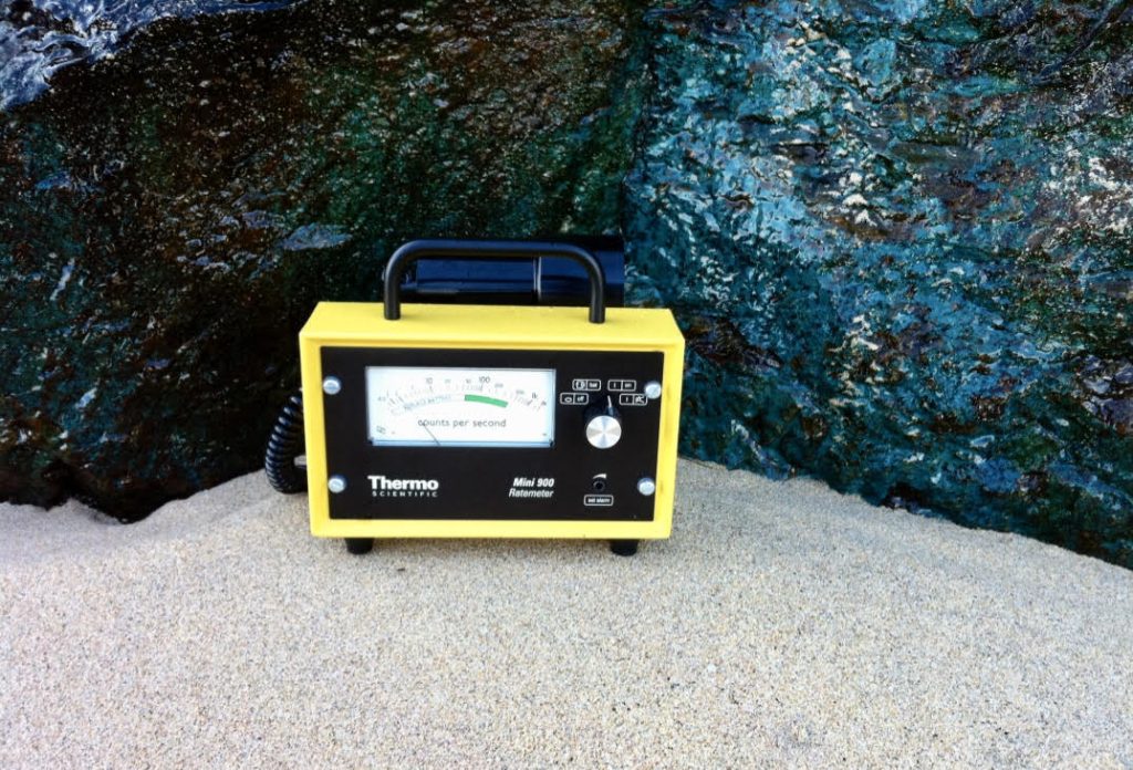 Image of a geiger counter, (a small device similar in design to a portable radio), placed on the sand in front of large rocks.