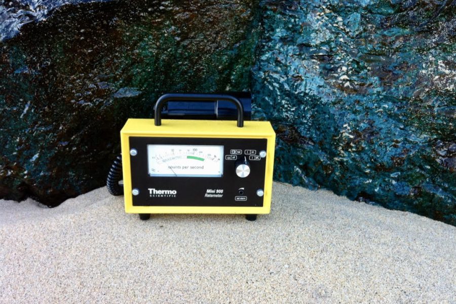 Image of a geiger counter, (a small device similar in design to a portable radio), placed on the sand in front of large rocks.